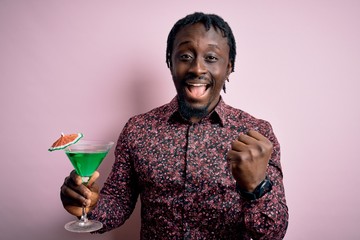 Poster - Young african american man drinking green cocktail. beverage over pink background screaming proud and celebrating victory and success very excited, cheering emotion