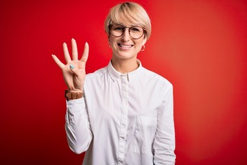 Wall Mural - Young blonde business woman with short hair wearing glasses over red background showing and pointing up with fingers number four while smiling confident and happy.