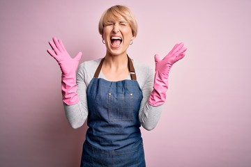 Sticker - Young blonde cleaner woman with short hair wearing apron and gloves over pink background celebrating mad and crazy for success with arms raised and closed eyes screaming excited. Winner concept