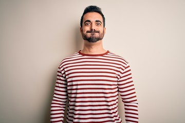 Sticker - Young handsome man with beard wearing casual striped t-shirt standing over white background puffing cheeks with funny face. Mouth inflated with air, crazy expression.