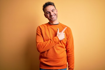 Young handsome man wearing orange casual sweater standing over isolated yellow background cheerful with a smile of face pointing with hand and finger up to the side with happy and natural expression