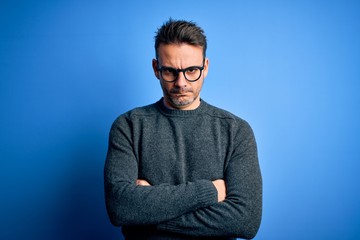 Young handsome man wearing casual sweater and glasses standing over blue background skeptic and nervous, disapproving expression on face with crossed arms. Negative person.