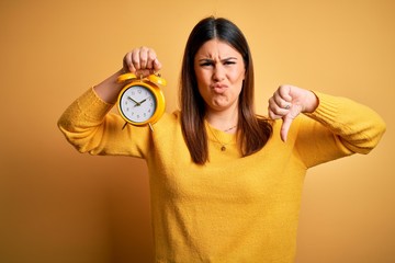 Poster - Young beautiful woman holding alarm clock standing over isolated yellow background with angry face, negative sign showing dislike with thumbs down, rejection concept