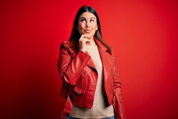 Sticker - Young beautiful woman wearing red leather jacket over isolated background with hand on chin thinking about question, pensive expression. Smiling with thoughtful face. Doubt concept.