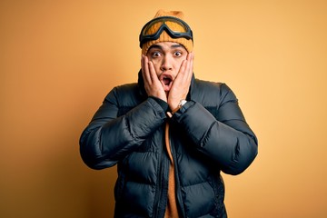 Wall Mural - Young brazilian skier man wearing snow sportswear and ski goggles over yellow background afraid and shocked, surprise and amazed expression with hands on face