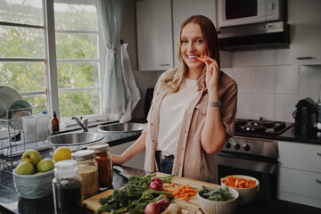 Wall Mural - Cheerful young woman in modern kitchen preparing fresh vegetables salad and eating a carrot smiling at the camera