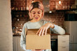 © shurkin_son - Cute young woman in casual clothes standing in kitchen interior with postal parcel, unpacking cardboard box full of groceries, having excited look. Home delivery, purchase and online shopping concept
