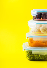 Stack of airtight glass food containers with colorful cooked vegetables on bright yellow background. Meal prep concept