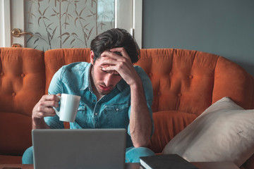 Handsome single man sitting on sofa in his house with coffee, looking in laptop and have headache