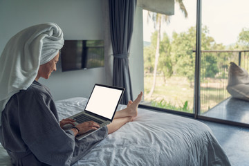 Wall Mural - Young beautiful young woman in a white shirt sits on a white bed in a hotel room and works on a laptop