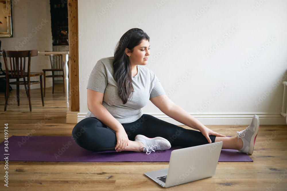 People, sports and fitness concept. Energetic plus size young woman in sneakers sitting on mat training, repeating exercises after professional instructor, watching online video lesson on laptop - obrazy, fototapety, plakaty 