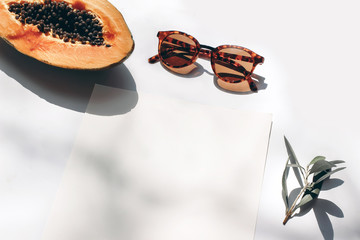 Wall Mural - Summer stationery still life scene. Closeup of cut papaya fruit, olive branch and sunglasses in sunlight. White table. Blank paper cards, invitations mockup scenewith harsh shadows. Top view.