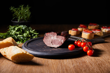 Wall Mural - selective focus of tasty ham on board near canape, parsley, cherry tomatoes and baguette on wooden table isolated on black