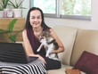 © Phuttharak - Asian woman sitting on couch in living room with  computer notebook  holding  Chihuahua dog  , looking and smiling at camera , work from home , stay home, stay safe concept.