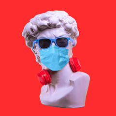 Statue. Earphone on a red background. Gypsum statue of David's head. Creative. Plaster statue of David's head in sunglasses and medical mask. Minimal concept art.