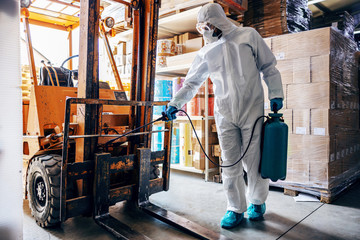 Wall Mural - Man in protective suit and mask disinfecting forklift in warehouse from corona virus / covid-19.