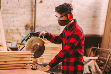 Wall Mural - Carpenter in protective glasses, headphones and mask using electric circular saw at sawmill. Professional skilled cabinet maker cutting wood board with circle saw. Woodworking, furniture production