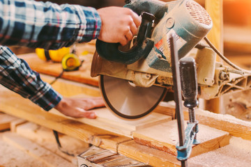 Wall Mural - Closeup of skilled man carpenter using electric circular saw at woodworking workshop. Professional cabinet maker cutting wooden board with circuit saw at sawmill. Wooden furniture production factory