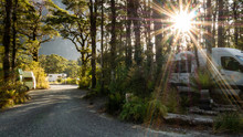 Sun Star Shot Of Campsite Located In Woods With Camper Vans, RVs And Gravel Path Leading Through Frame. Photo Taken In Milford Sound, Fiordland National Park, New Zealand