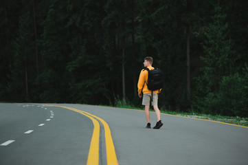 Wall Mural - Young man tourist in casual clothes and backpack walking along a forest road and looking around with a serious face. Stylish guy walking on a mountain asphalt road.