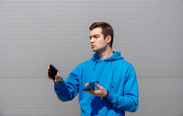 young man with earring holds two smartphones in both hands and look at them with serious face, wearing in blue hoodie and black fitness bracelet, standing against the gray textured wall