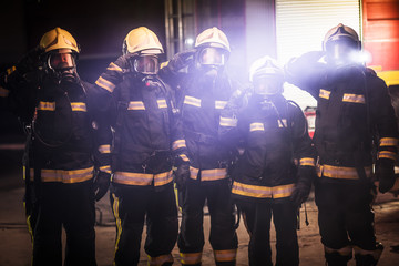 Wall Mural - Group of professional firefighters posing. Firemen wearing uniforms, protective helmets and oxygen masks. Smoke and firetrucks in the background.