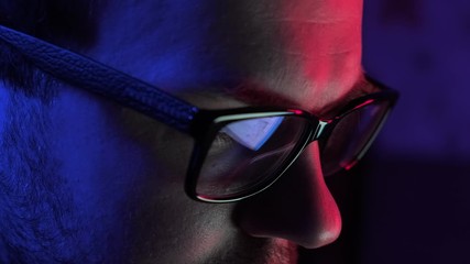 Male hacker coding and programming using laptop. Close-up of man in glasses hacking on computer. Blue and red lights are reflected on him. Cybercrime concept. 4K, UHD