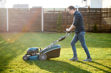 Spring Mowing The Lawn. Young Man In A Plaid Shirt And Jeans Mows Grass With A Lawn Mower In The Backyard. Side View