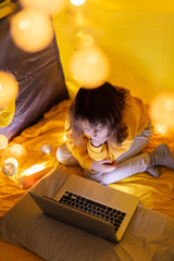 Wall Mural - young girl student using laptop for homeschooling in a home made tent with glowing light balls.