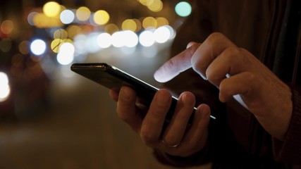 Male hands typing text message in social network chats via cell mobile phone at night urban lights on background. Blurred background and bokeh effects.