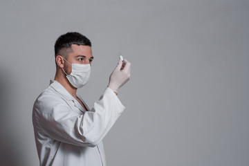 A man in a white medical gown, a mask on his face and surgical gloves looks at a glass jar of vaccine on a white or gray background..