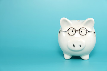 White ceramic piggy bank wearing reading glasses on blue teal background. Concept for money savings plan for retirement, aged society, or financial accounting