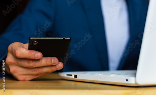 Close-up of the hands of a well-dressed man in a blue suit and a white shirt looking at his smartphone, using a laptop on a desk Poster Mural XXL