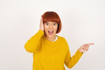 Wall Mural - Portrait of young woman with shocked facial expression, showing something amazing on blank space, one hand on her head and pointing with forefinger. OMG concept.