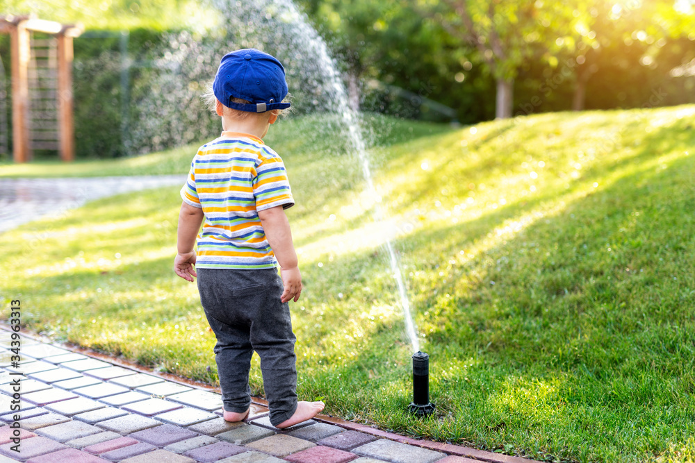 Cute adorable caucasian blond barefeet toddler boy in cap walking at home backyard near sprinkler automatic watering system lawn in garden. Child little helper playing gardening at summer outdoors - obrazy, fototapety, plakaty 