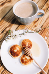 Sticker - Delicious three cottage cheese pancakes with honey lying on the white plate with the branch of a flowering tree on the wooden table background with the cuo of coffee. Curd cheese fritters.