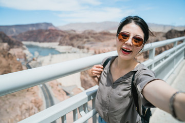 delightful female tourist taking selfie with background hoover dam on sunny day. happy summer vacation chinese girl visiting and taking photo by handrail with famous landmark reservoir united states.