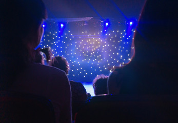 Blurred view of audience back. Silhouette shot from back in blue light. Multicolored light beams spotlights at the time of the entertainment show. Blurred glowing lights on stage. Small depth of view.