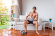 © wirojsid - Attractive handsome muscular man doing dumbbell weight exercises in living room at home, sitting on sofa