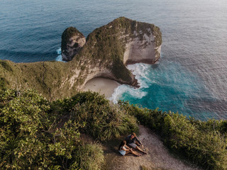 Couple sitting on the clif and looking on popular place in Nusa Penida - Kelingking beach. Travelling couple. Aerial view of the small island of Nusa Penida Island