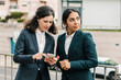 © Mangostar - Concentrated businesswomen using smartphone. Female colleagues in formal wear standing on street and using cell phone together. Business and technology concept