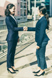 © Mangostar - Smiling businesswomen shaking hands. Side view of cheerful female colleagues in formal wear standing on street and greeting each other. Partnership concept