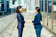 © Mangostar - Content businesswomen talking on street. Side view of cheerful female colleagues standing outdoors, shaking hands and talking. Handshake concept