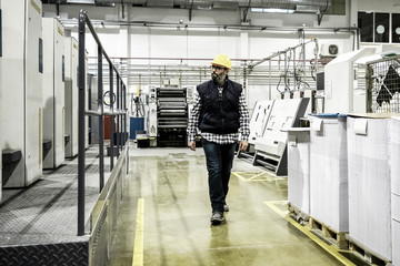 Wall Mural - Confident printing worker walking at factory. Mature man in yellow hard hat walking near printing machine and looking aside. Print manufacturing concept