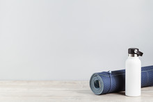 Rolled Blue Yoga Mat And White Metal Water Bottle Flask On Grey Wooden Surface. Gender Neutral Fitness And Exercise Concept And Hydration With Copy Space. Active Lifestyle. Workout At Home