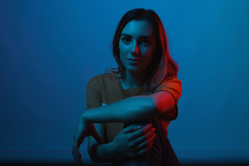 Red and blue light portrait of a young woman sitting with arm on a knee in a studio.