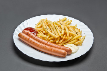 White ceramic plate with French fries, two grill sausages, ketchup and mayonnaise on grey background