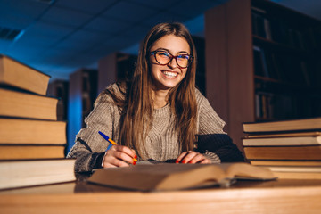 Young happy smiling student in glasses preparing for the exam. Girl in the evening sits at a table in the library with a pile of books, smiling and looking at the camera