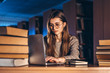 © bedya - Young student woman in the evening sits at a table in the library with a pile of books and works on a laptop. Preparing for the exam