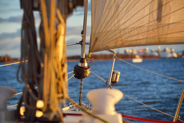 White yacht sailing in Daugava river at sunset. Close-up view from the deck to the bow, mast and sails. Cargo ships and port cranes in the background. Riga bay, Latvia